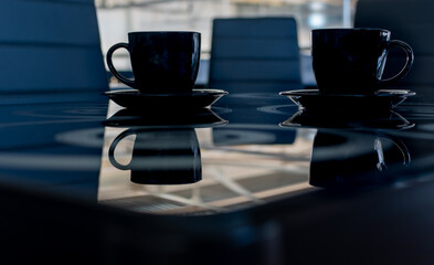 Black cups for tea or coffee on black glass table with reflections on blurred background of office soft chairs