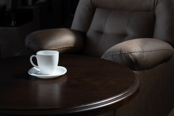 White teacup on round wooden table on background of luxurious soft leather chair in soft sun light