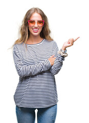 Beautiful young blonde woman wearing sunglasses over isolated background with a big smile on face, pointing with hand and finger to the side looking at the camera.