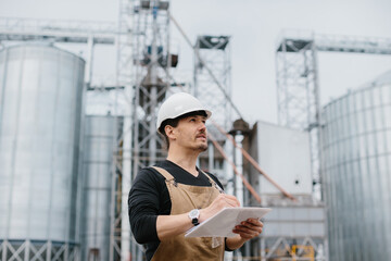 A young engineer in a hard hat with a project in his hands checks the status of the silo construction. © dsheremeta