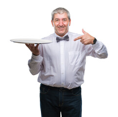 Handsome senior waiter man holding silver tray over isolated background with surprise face pointing...