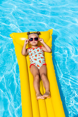 Cute little girl lying on inflatable mattress in swimming pool with blue water on warm summer day...