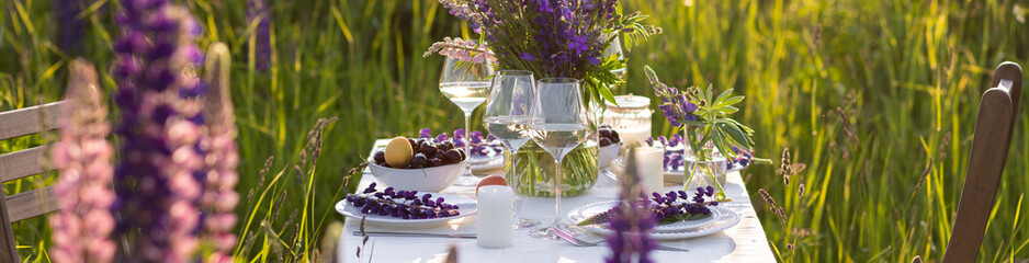 Beautiful countryside wedding of romantic dinner decor on blooming meadow with purple violet lupins flowers. Vase, wineglasses, cherry, apricot. Wooden table, basket. Sunset golden hour, summer banner