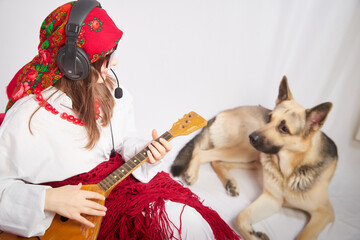 A girl in red scarf, white shirt, with shawl and balalaika in black sunglasses with a shepherd dog. A cheerful radio presenter with headphones and a microphone sings and has fun with an animal friend