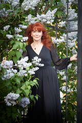 Elegant senior mature Woman in Black Dress by Blooming Lilac Bush at Dusk. A woman with red hair stands poised among lilac blooms