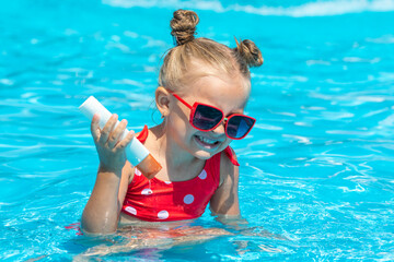 cute little girl in sunglasses and swimsuit has fun in the pool and holding sunscreen for face. summer vacation of the child, protection of children's skin from the sun