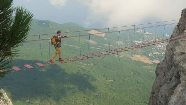 Woman backpacker walking on suspension bridge in mountains over precipice.