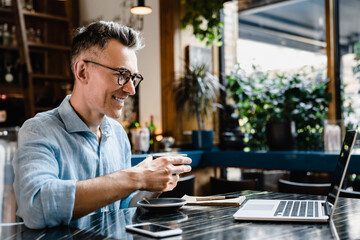 Confident successful caucasian mature businessman freelancer boss self-emloyed enjoying morning breakfast coffee in cafe restaurant working remotely on laptop, watching webinars online
