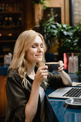 Having breakfast out. Young caucasian businesswoman freelancer boss self-employed drinking coffee while working on laptop remotely on distance, smelling coffee aroma at the bar counter.