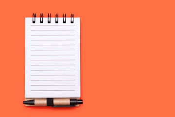 Spiral paper notebook with small pen on orange background with copy space, place for text. A small lined notebook with free space for notes. Open paper notebook with blank white lined pages