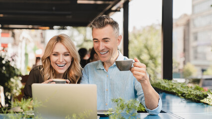 Laughing smiling caucasian middle-aged couple spouses friends colleagues watching movie webinar together on laptop while on a date in cafe restaurant