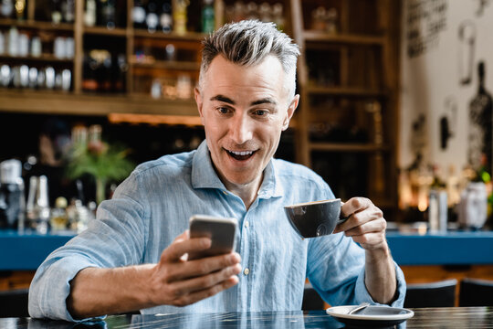 Shocked impressed caucasian middle-aged mature businessman freelancer boss self-employed man using smartphone, reading news, e-banking, loan, lottery winner in cafe restaurant