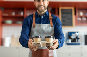Fototapeta na wymiar Smiling male barista in apron holding two disposable cups of coffee, working in cafe. Takeaway coffee concept