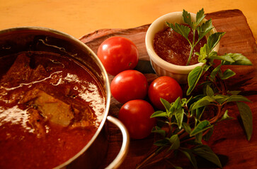 A small pan with beef tongue in sauce, cherry tomatoes, basil and a container with pepper sauce and mint sprigs.