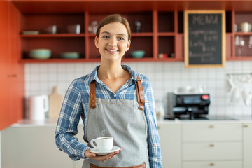 Cheerful female barista holding cup of coffee and smiling to camera, standing at her own coffee...