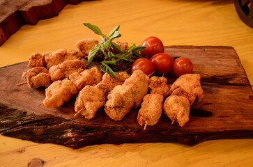A wooden board with breaded chicken skewers, cherry tomatoes and sprigs of basil and rosemary.