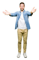 Handsome man with blue eyes and beard wearing denim jacket looking at the camera smiling with open arms for hug. Cheerful expression embracing happiness.