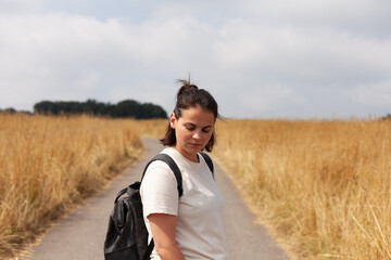 Young woman with a rucksack on a rural road, surrounded by the beauty of a golden wheat field and...