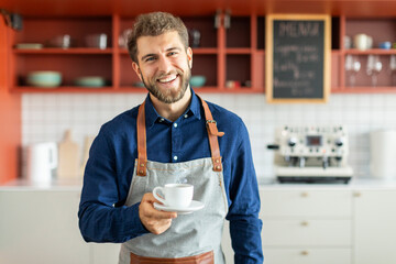 Handsome bearded barista man small business owner holding cup of coffee and smiling, standing in...