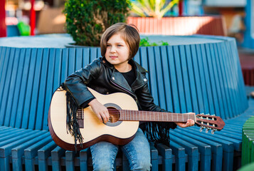 A beautiful little girl, in rock style,in a leather jacket and jeans,sits and plays an acoustic...