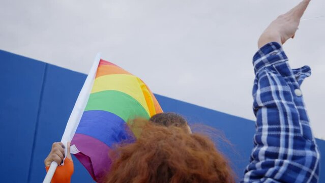 Young smiling drag queen waving rainbow flag on gay pride festival day in front of a blue wall. Community of happy transgenders people celebrating LGBT free love and sex festival
