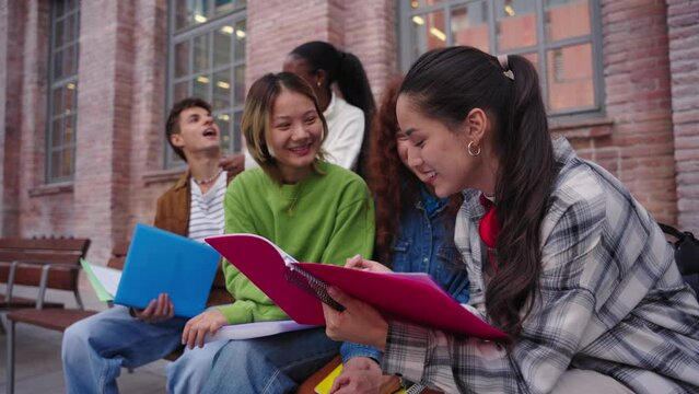 Smiling group of young students sitting on bench studying together with workbooks outside university building. Academics multiracial people talking and having a good time gathered on the campus