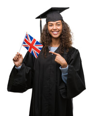 Young hispanic woman wearing graduation uniform holding flag of UK very happy pointing with hand...