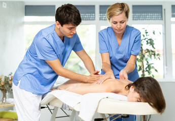 Obraz premium Young masseur trainee practicing skills with experienced woman massage therapist guiding him during back massage session for female patient..