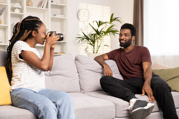 Obraz premium Happy African American Couple Enjoying Photography At Home. Cheerful Woman Taking Pictures Of Man With Digital Camera. Leisure Activities, Hobbies, And Modern Lifestyle Concept In Cozy Living Space.