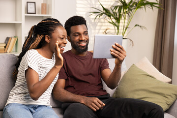 Happy Couple Enjoying Technology On Tablet At Home. A Smiling Man And Woman Sit On Couch, Sharing A Fun Moment While Looking At A Digital Screen. Lifestyle Concepts In Modern Interior Setting.