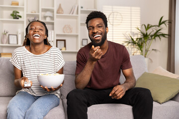 Joyful African American Couple Relaxing On Sofa With Popcorn, Happy Moments Together, Indoor Leisure And Comfort, Smiling Man And Woman Watching TV
