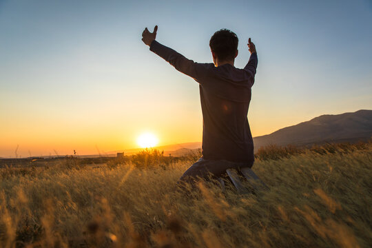 Boy kneeling praying with arms outstretched at sunset, spiritual concept, stock photo