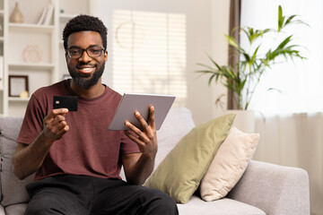 Smiling Man With Credit Card And Tablet At Home. Cheerful Young African American Male Holding Payment Card, Online Shopping, Indoor Comfort, Digital Economy And Convenient Banking.
