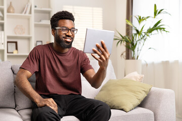 Happy African American Man Using Tablet On Couch At Home. Smiling Male Engaged In Video Call, Leisure Tech Lifestyle. Comfortable Living and Modern Connectivity Concept.