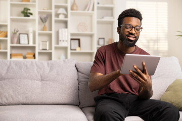 Smiling African American Man Using Tablet On Couch At Home. Casual Lifestyle, Digital Literacy, E-Learning Concept. Joyful Male Engaged In Social Media Or Working Remotely. 