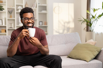 Happy Man Enjoying Coffee On Couch At Home. Relaxed Adult With Glasses Holding Mug, Smiling In Cozy Living Room Interior. Casual Lifestyle, Comfort, Leisure Concept.