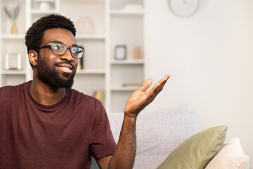 Happy African American Man Sitting On Couch At Home With Hand Gesture. Casual Comfort, Smiling, Relaxing Lifestyle Concept. Cozy Modern Living Room Interior.