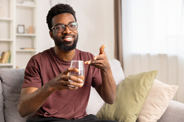 Happy African American Man Enjoying Drink At Home. Confident Male With Casual Wear, Smiling And Holding Glass Of Water. Comfort, Hydration, Lifestyle And Wellness Concept