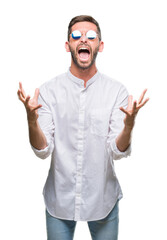 Young handsome man wearing glasses over isolated background crazy and mad shouting and yelling with aggressive expression and arms raised. Frustration concept.