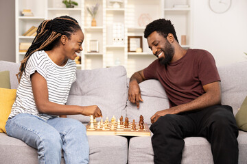 Happy Couple Enjoying A Game Of Chess At Home. Smiling African-American Man And Woman Engaged In A Friendly Chess Match, Experiencing Joy And Leisure In Comfortable Living Room Setting.