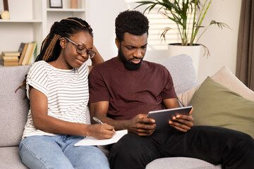 Couple Planning Together, African American Man And Woman Writing Notes And Using Tablet On Sofa At Home. Concept Of Teamwork, Collaboration, Home Budget Planning. Diverse, Happy, Technology