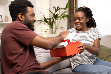 A Joyful African American Couple Sharing A Gift Box While Sitting On A Sofa, Embodying Love, Generosity, And Togetherness.