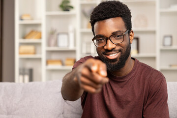Smiling African American Man Pointing At Camera In Modern Living Room. Selective Focus On Face With Blurry Bookshelf Background. Concept Of Invitation, Engagement, And Direct Communication