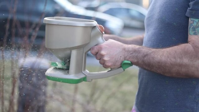 Man Fertilizing Lawn with Handheld Spreader. Close-up of a man's hands using a manual seed spreader to fertilize a grass lawn.