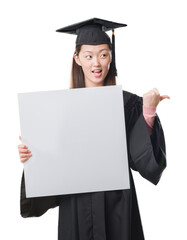 Young Chinese woman wearing graduate uniform holding banner pointing and showing with thumb up to...