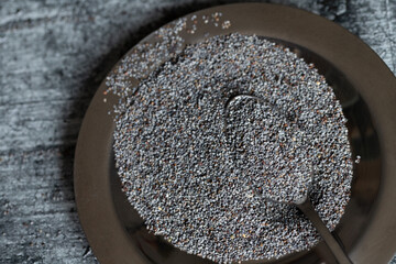 Close-up of a metal bowl containing blue poppy seeds. A spoon is in the poppy seed. The background is gray. - obrazy, fototapety, plakaty