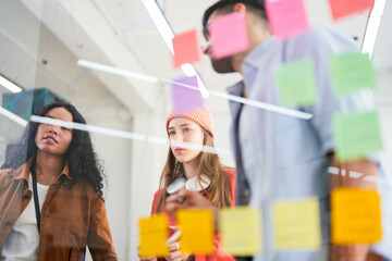 Business team uses a glass wall covered with sticky notes to outline processes during meeting. Engaged colleagues gather around a glass brainstorming board. plotting ideas and strategies.