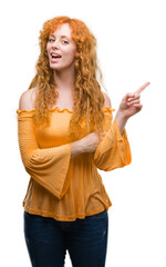 Young redhead woman with a big smile on face, pointing with hand and finger to the side looking at the camera.