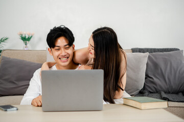 Young couple is sharing a cheerful moment on a couch with a laptop. The man, dressed in a white...