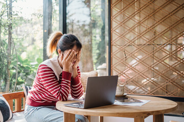 Overwhelmed young woman covering her face, seated before a laptop in a well-lit home office setting.